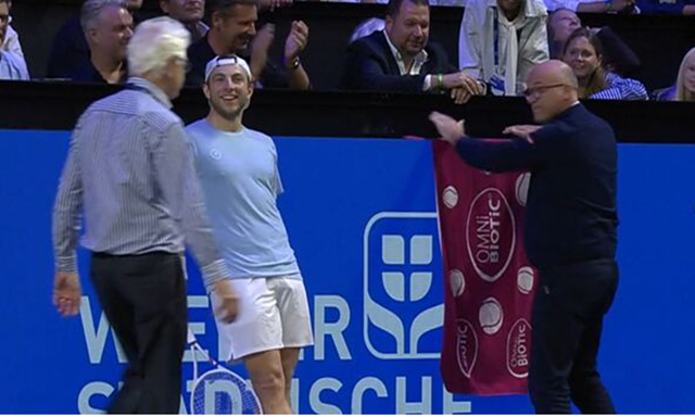 Tallon Griekspoor (center) smiles as he watches an official cover the malfunctioning advertising board with a towel during the match. On the right is the official covering the advertising board with a red towel. /Photo=Tennis TV capture