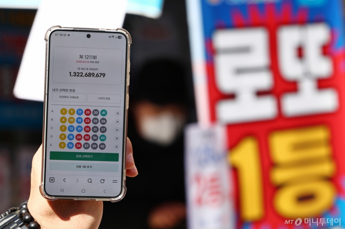 24년 만에 모바일 로또 판매가 시작된 9일 오전 서울 시내 한 복권 판매점 앞에서 휴대폰 화면에 나오는 로또 모바일 판매 페이지 뒤로 시민들이 로또를 구매하고 있다. 이용자는 모바일 동행복권 홈페이지에 가입한 뒤 예치금을 충전하면 오전 6시부터 자정까지 복권을 살 수 있다. 올해 상반기에는 평일에만 회차당 1인 5000원까지 살 수 있으며, PC 온라인 구매 금액과 합산해 계산된다. 토요일에는 모바일 구매가 막힌다. 전체 온라인 판매 규모도 전년도 로또 판매액의 5% 이내로 묶여, 한도에 도달하면 판매가 자동 중단된다. /사진=뉴시스