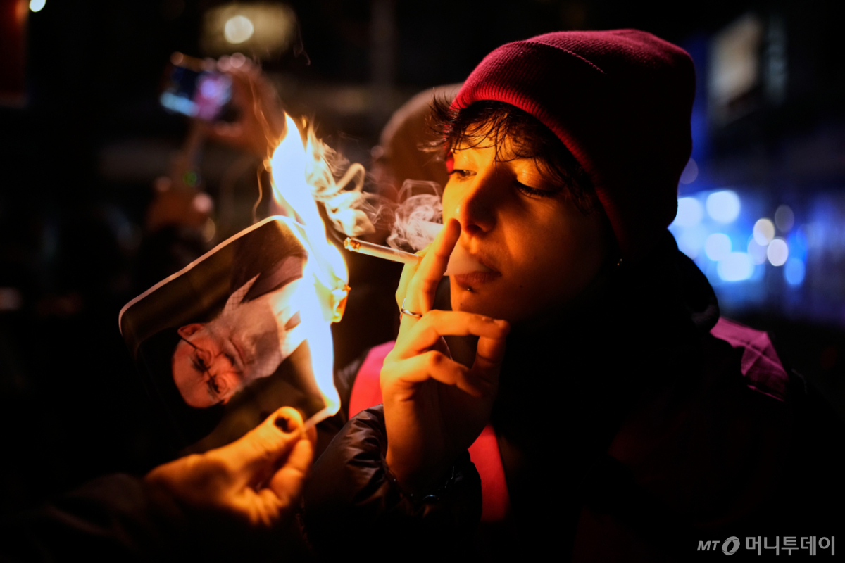 이란의 대규모 반정부 시위를 지지하는 한 시위자가 지난 1월 14일 독일 베를린에서 이란 최고 지도자 아야툴라 알리 하메네시의 포스터에 붙인 불로 담배불을 붙여 담배를 피우고 있다. 자신을 캐나다에 거주하는 이란 난민이라고 소개한 한 여성이 이란 최고지도자 아야툴라 알리 하메네이의 사진에 라이터로 불을 붙이고 그것으로 다시 담배에 불을 붙여 한 모금 빨아들인 뒤 담배를 버리는 모습으로 전 세계에서 이란 저항의 상징으로 떠올랐다./AP=뉴시스